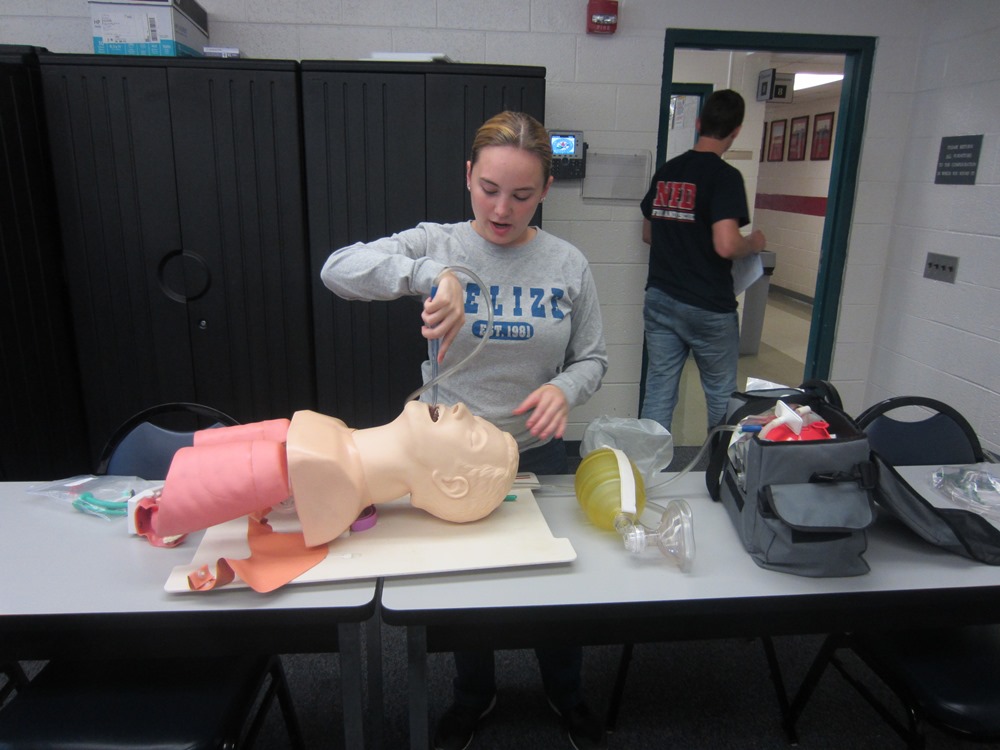 Photo of an EMT student practicing emergency techniques with a dummy lying on the table and another person walking out of the classroom